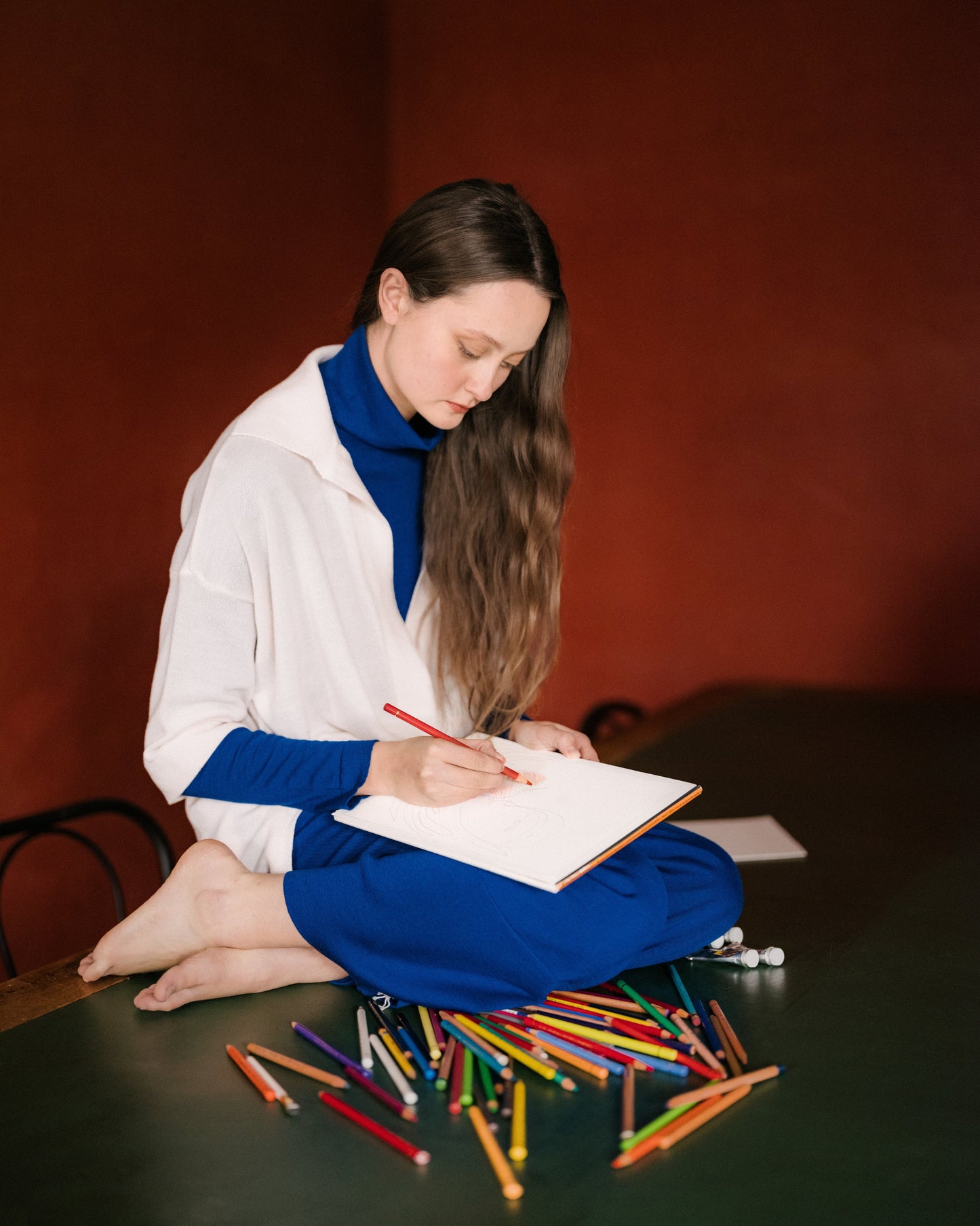 Model seated on a table sketching with colored pencils, wearing a white maxi polo over a blue outfit