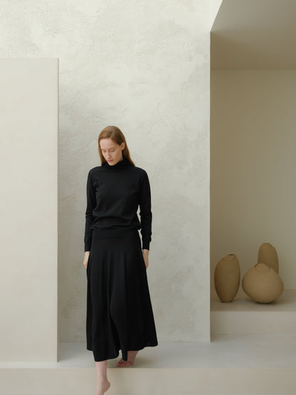 Model wearing a black turtleneck and ankle-length 3D-knit Extra Fine Merino Wool A-Line Skirt in a minimalist studio.