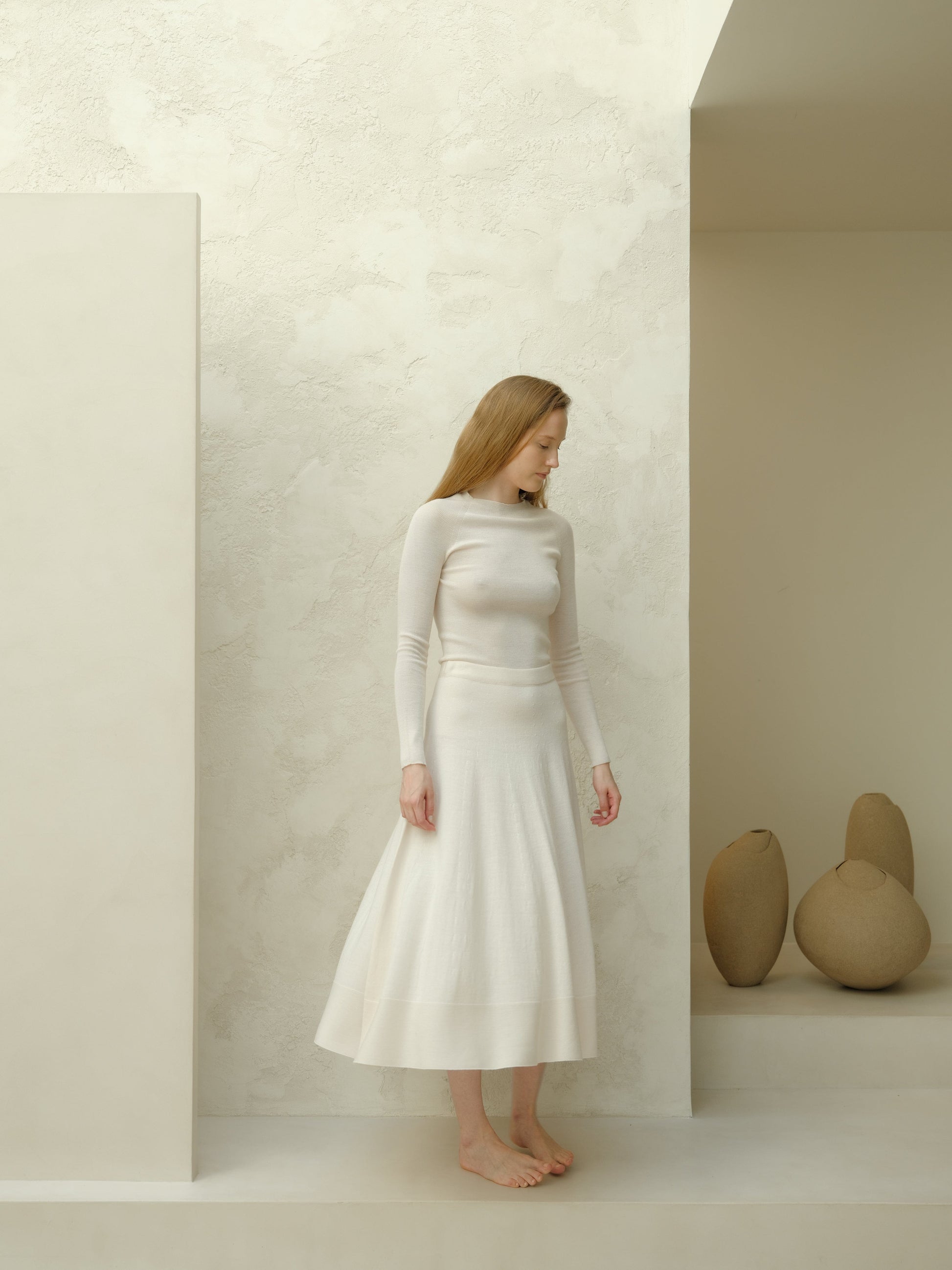 Model wearing ivory long sleeve knit top and white merino wool 3D-knit A-line skirt in a minimalist studio.