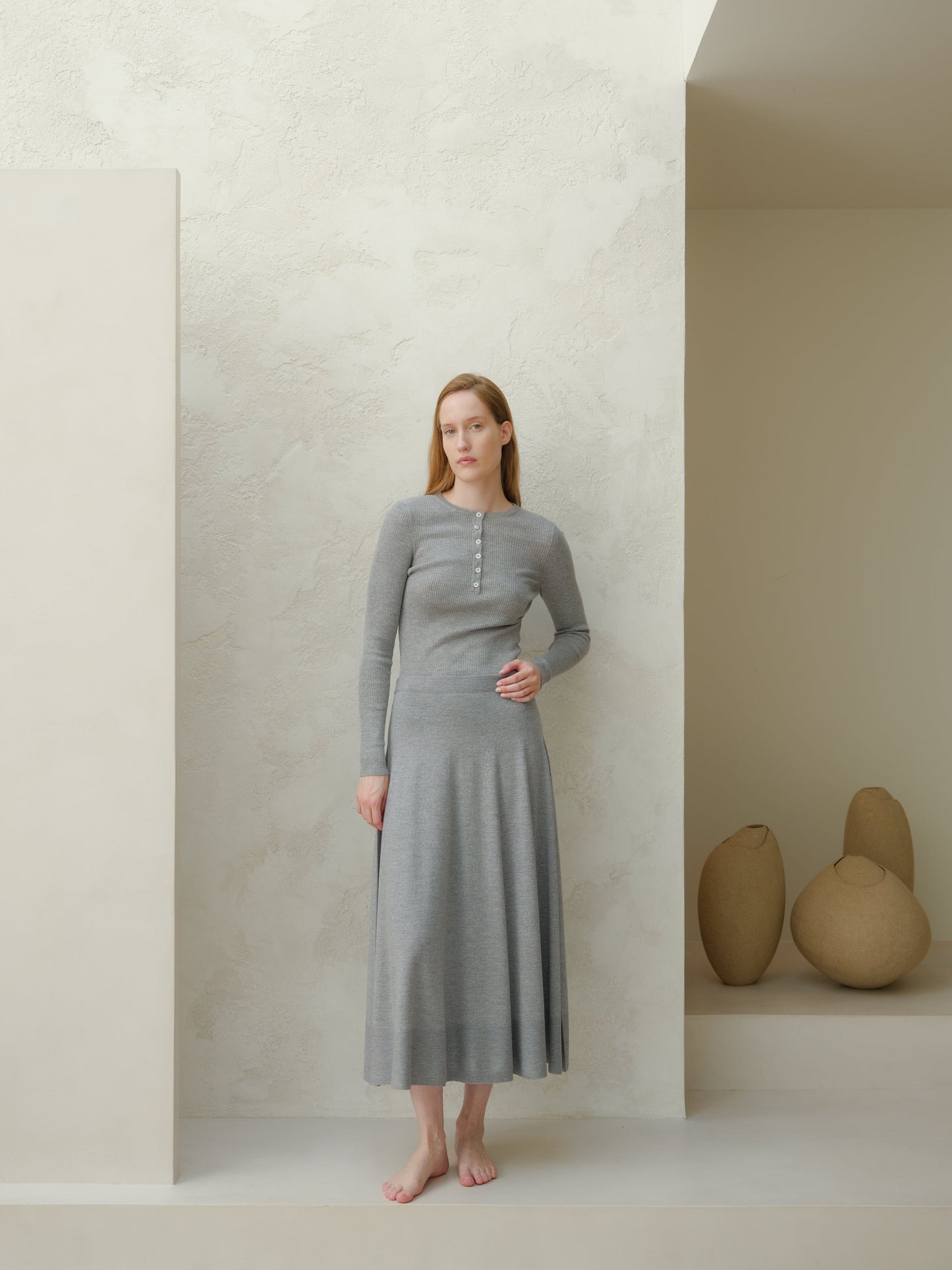 Front view of a model wearing a gray merino wool A-line skirt with a matching knit top, barefoot against a textured wall.