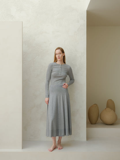 Front view of a model wearing a gray merino wool A-line skirt with a matching knit top, barefoot against a textured wall.