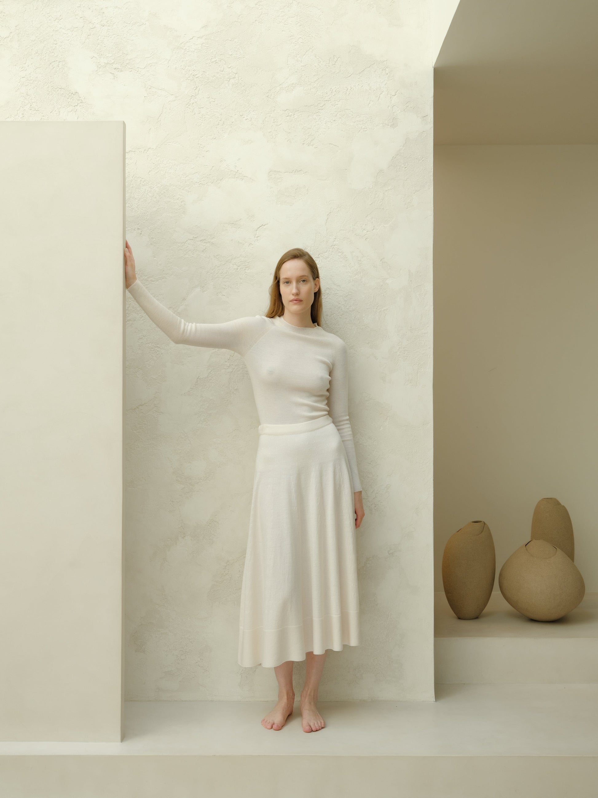 Ivory 3D-knit merino wool A-line midi skirt with a matching knit top, model standing barefoot against a pale textured wall.