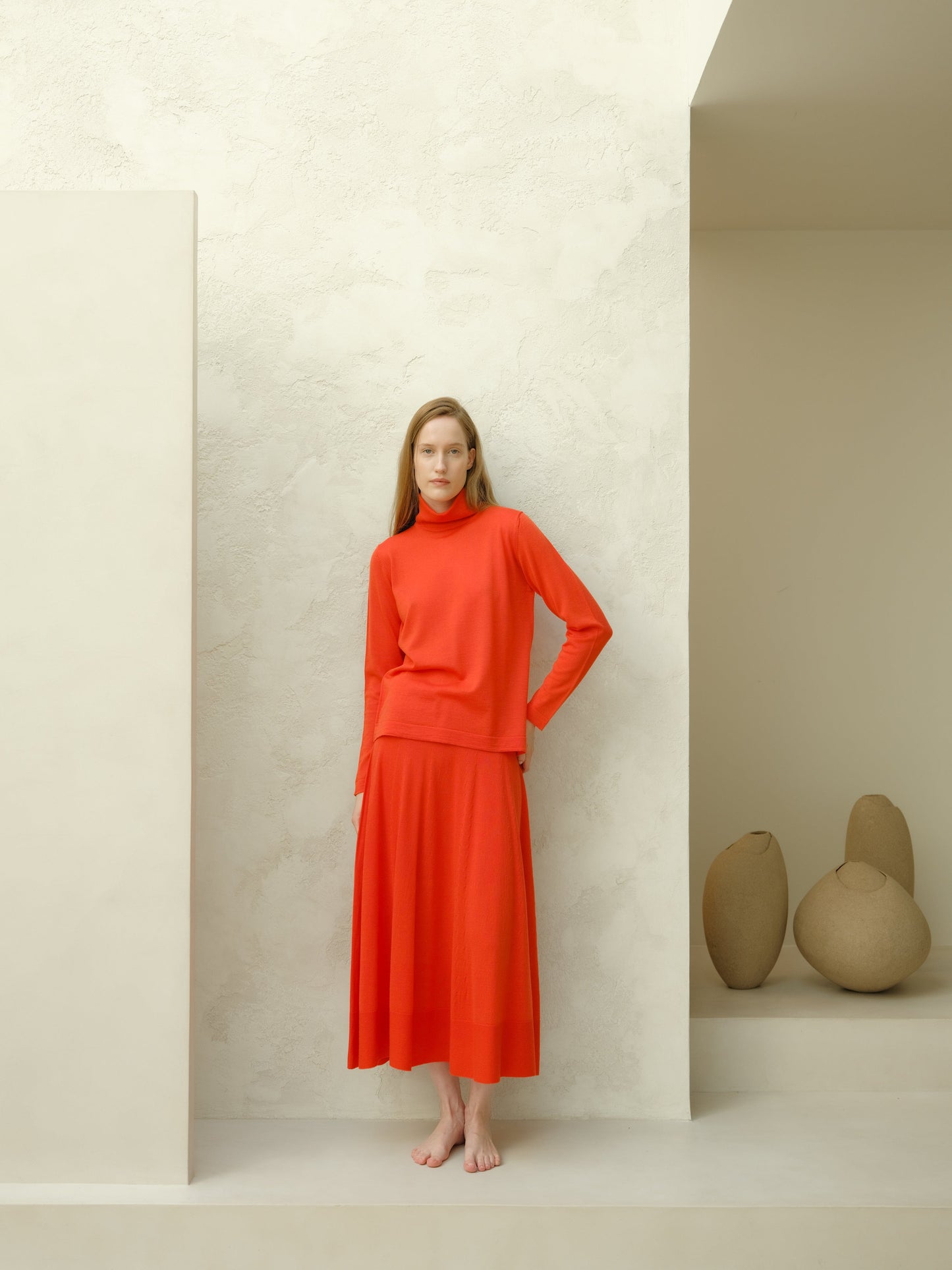 Model in coral red merino wool turtleneck and long A-line skirt stands barefoot in a pale studio with rounded vases on a ledge.
