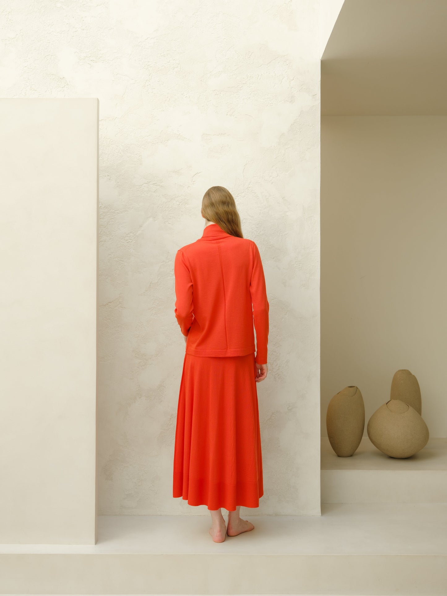 Rear view of a model in a bright orange merino wool A-line skirt with a matching top standing barefoot.