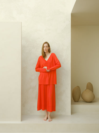 Model wearing coral red merino wool V-neck sweater and long A-line skirt in a minimalist pale studio setting.
