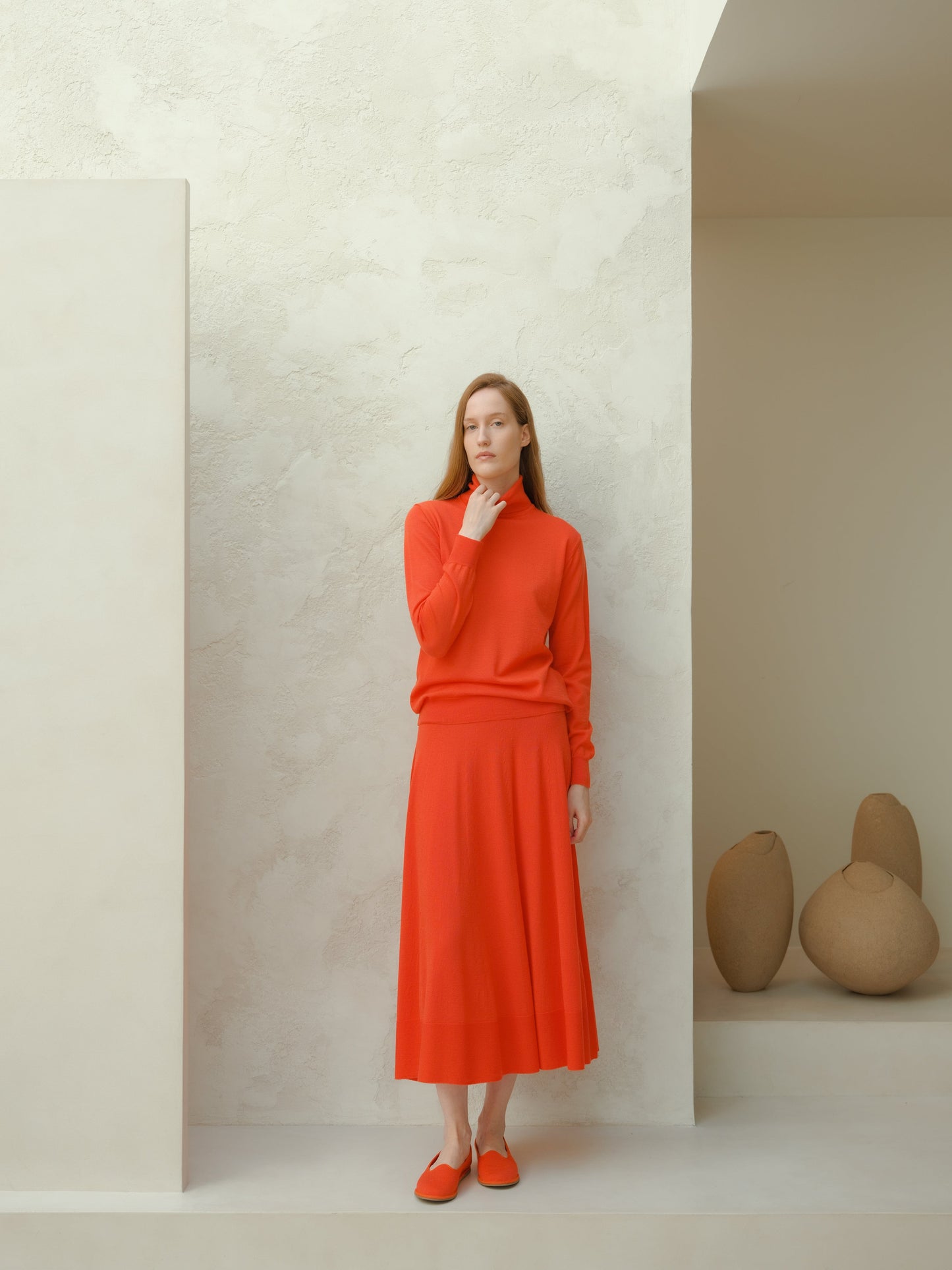Model in coral red merino wool turtleneck and long A-line skirt stands against a textured white wall, orange flats completing the look.