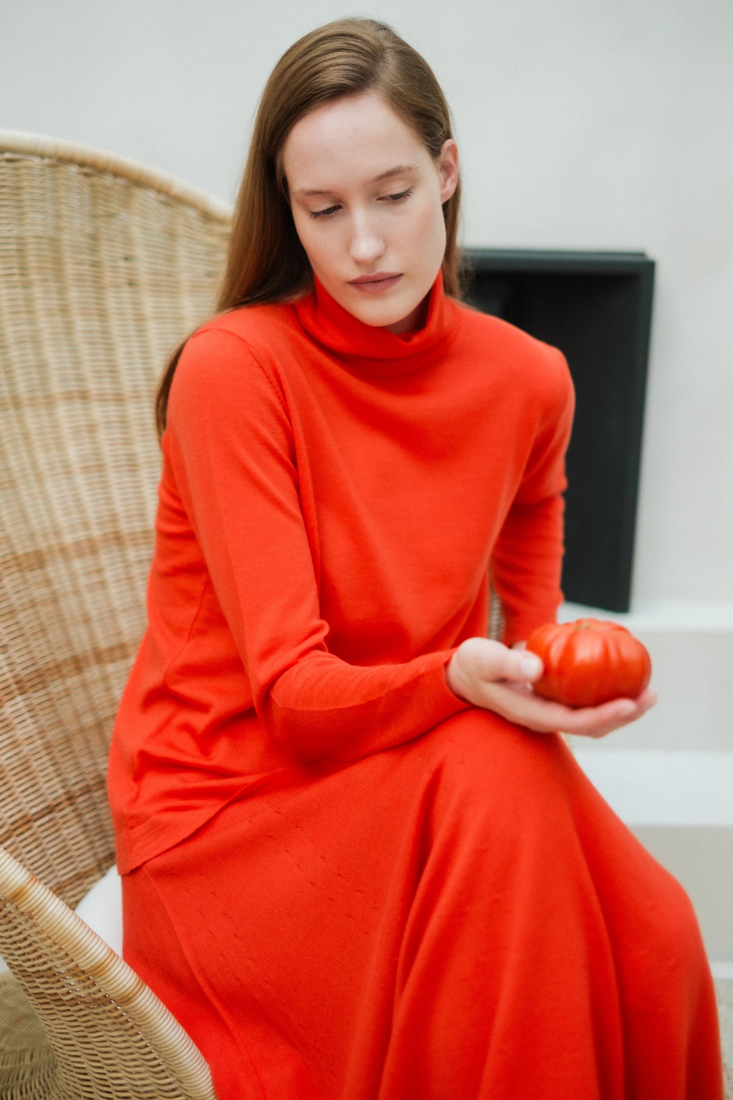 Woman in bright orange knit turtleneck and long A-line skirt sits on a wicker chair.