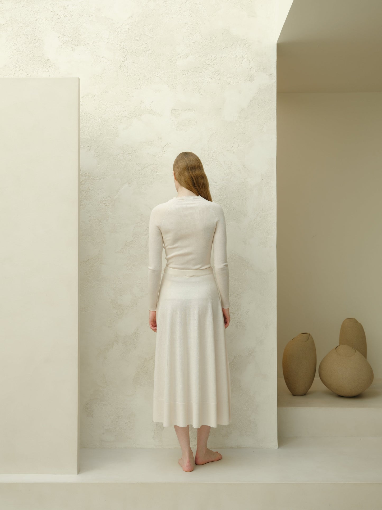 Back view of ivory 3D-knit merino wool A-line midi skirt with a matching knit top, standing barefoot in a cream studio.