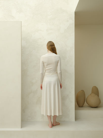 Back view of ivory 3D-knit merino wool A-line midi skirt with a matching knit top, standing barefoot in a cream studio.