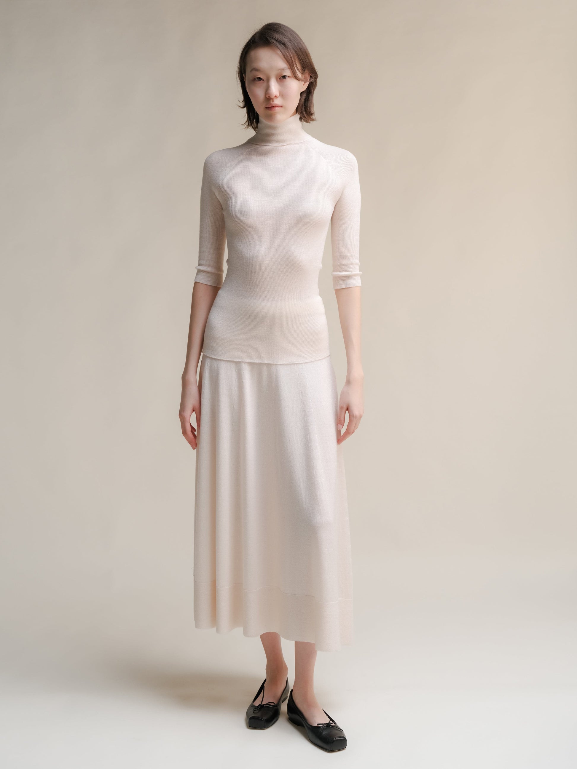 Full-length view of a model in ivory 3D-knit merino wool A-line skirt with a matching turtleneck and black flats in a neutral studio.