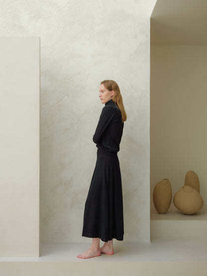 Side view of a model wearing a black 3D-knit merino wool A-line skirt with a black turtleneck