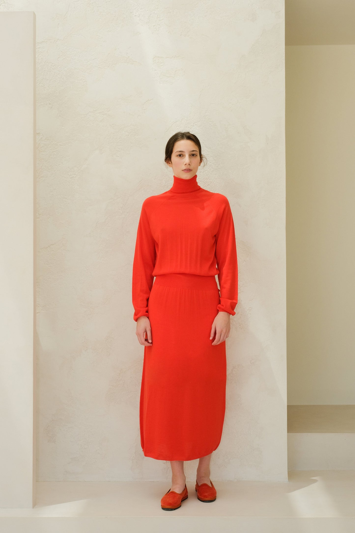 Model facing forward in a bright red 3D knit merino wool dress with a high neck and ribbed waist, midi-length.