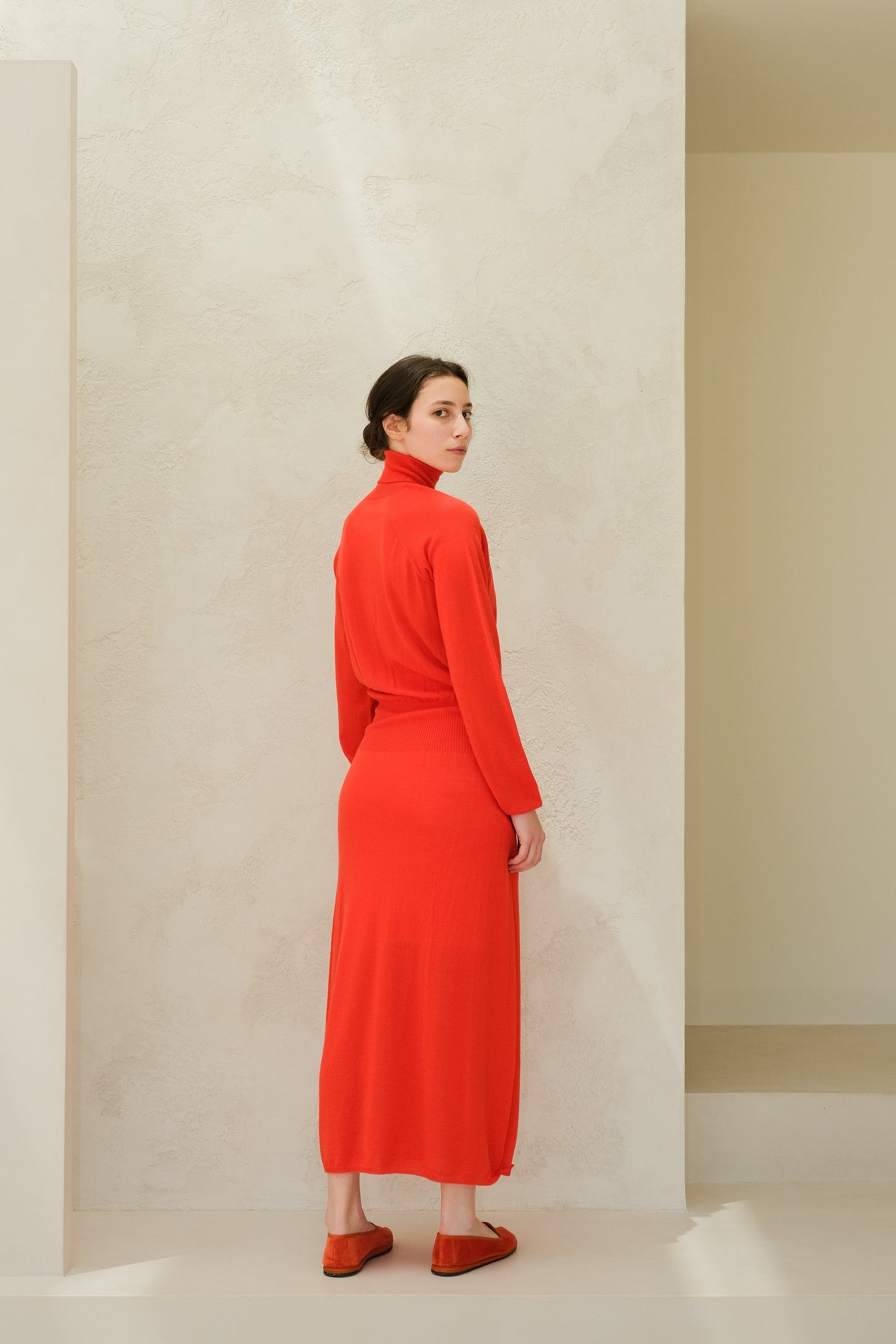Model wearing bright red merino wool 3D-knit dress with a turtleneck, seen from the back.