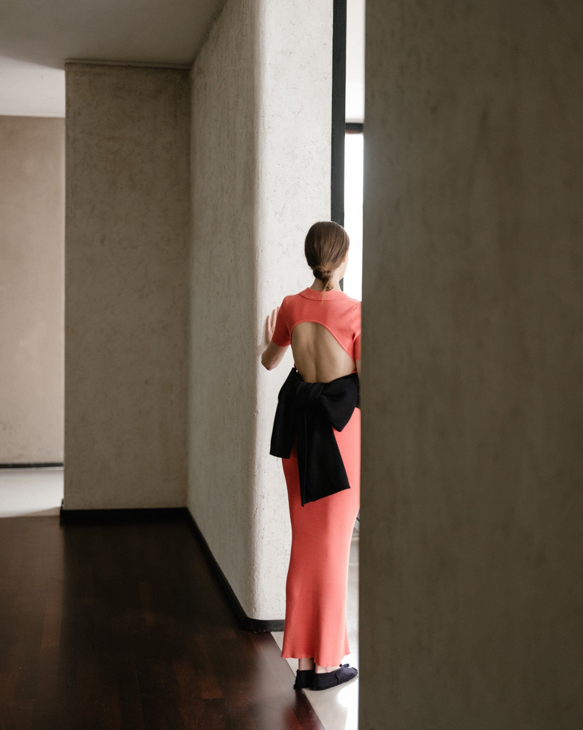 Back view of a model in a coral open-back dress with an oversized black bow tied at the lower back.