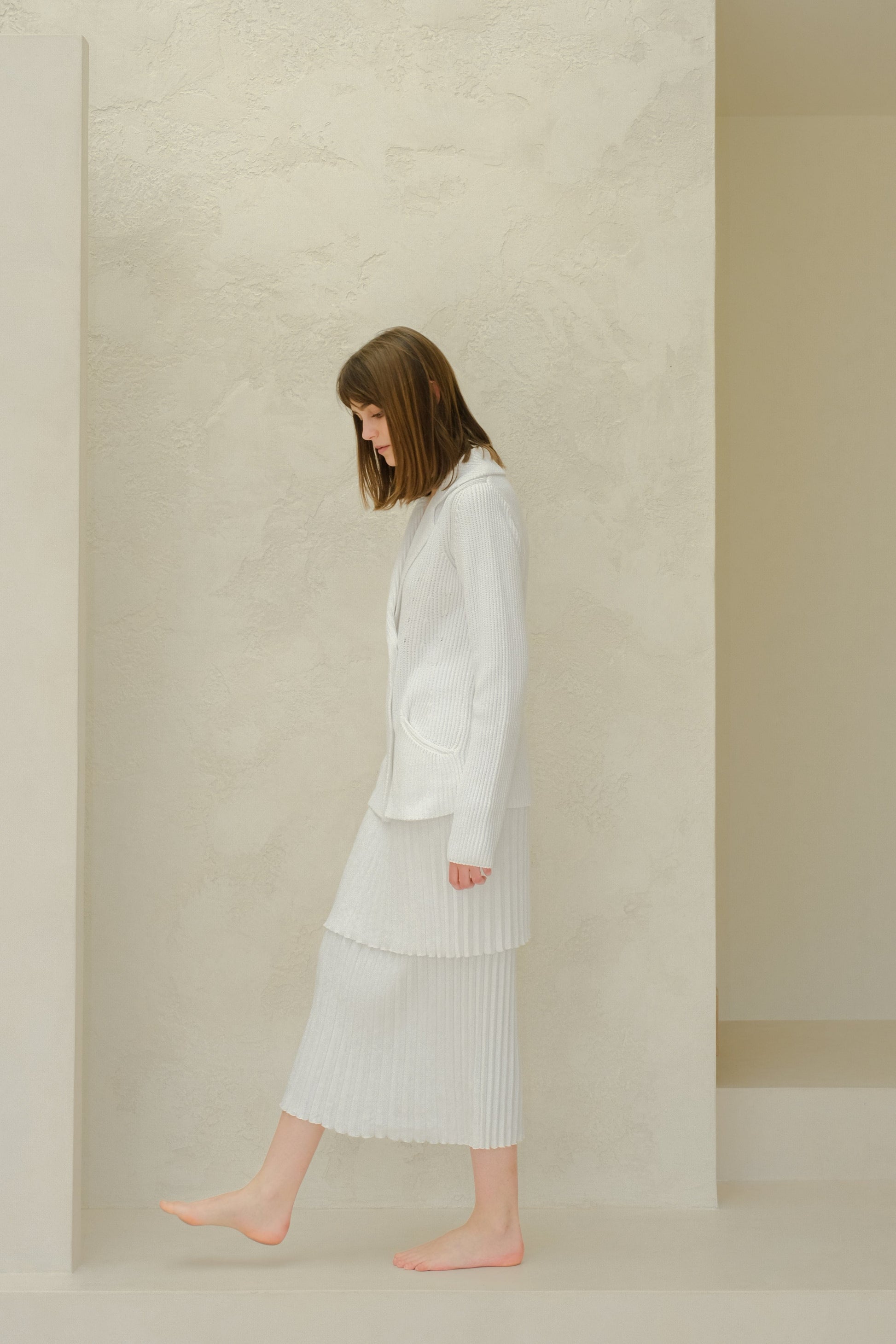 Side profile of a model in a white knitted blazer with a layered pleated long skirt from Fine Cotton Plissé