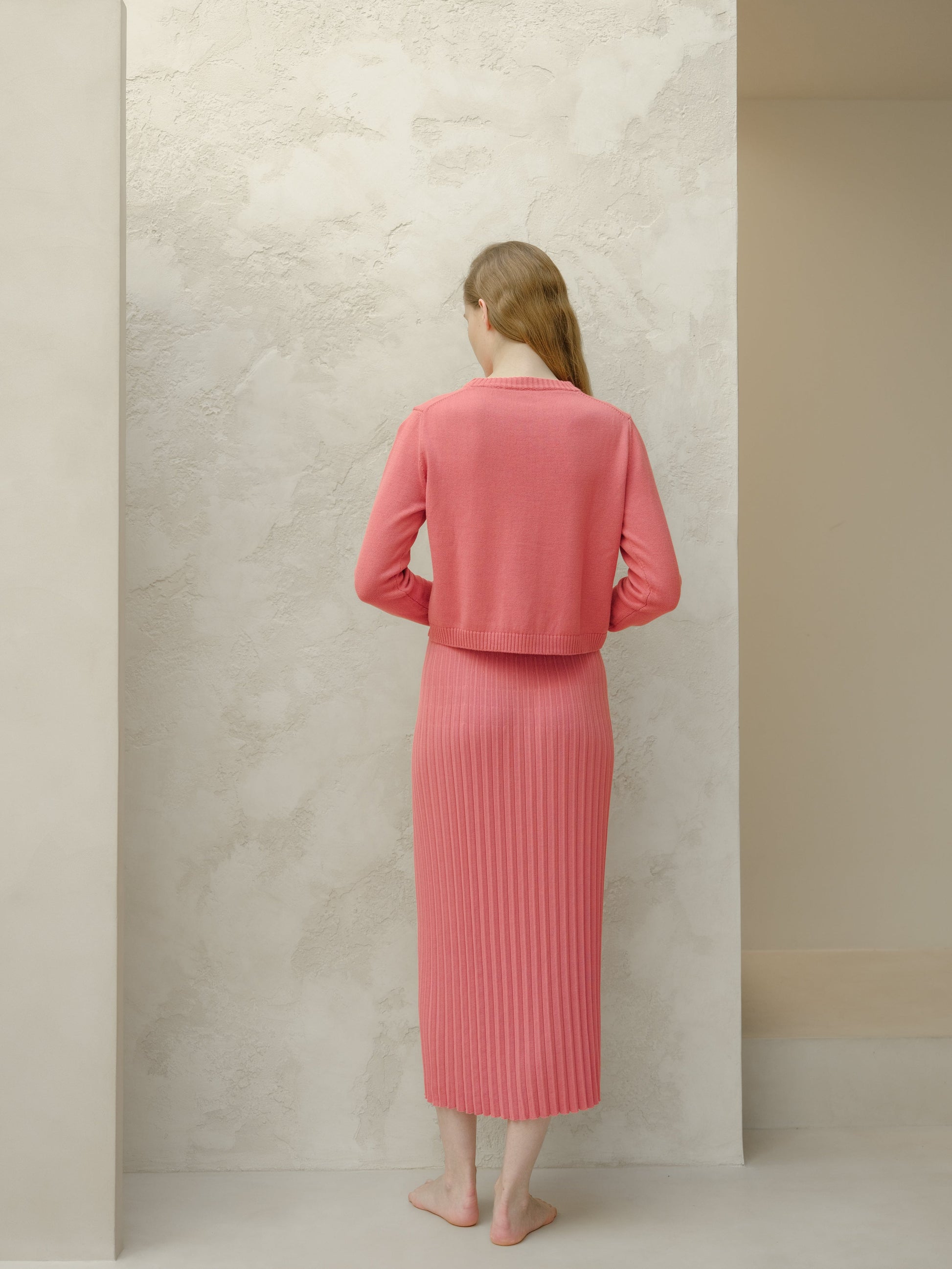 Back view of coral knit cropped cardigan with a matching long pleated skirt worn by a barefoot model.