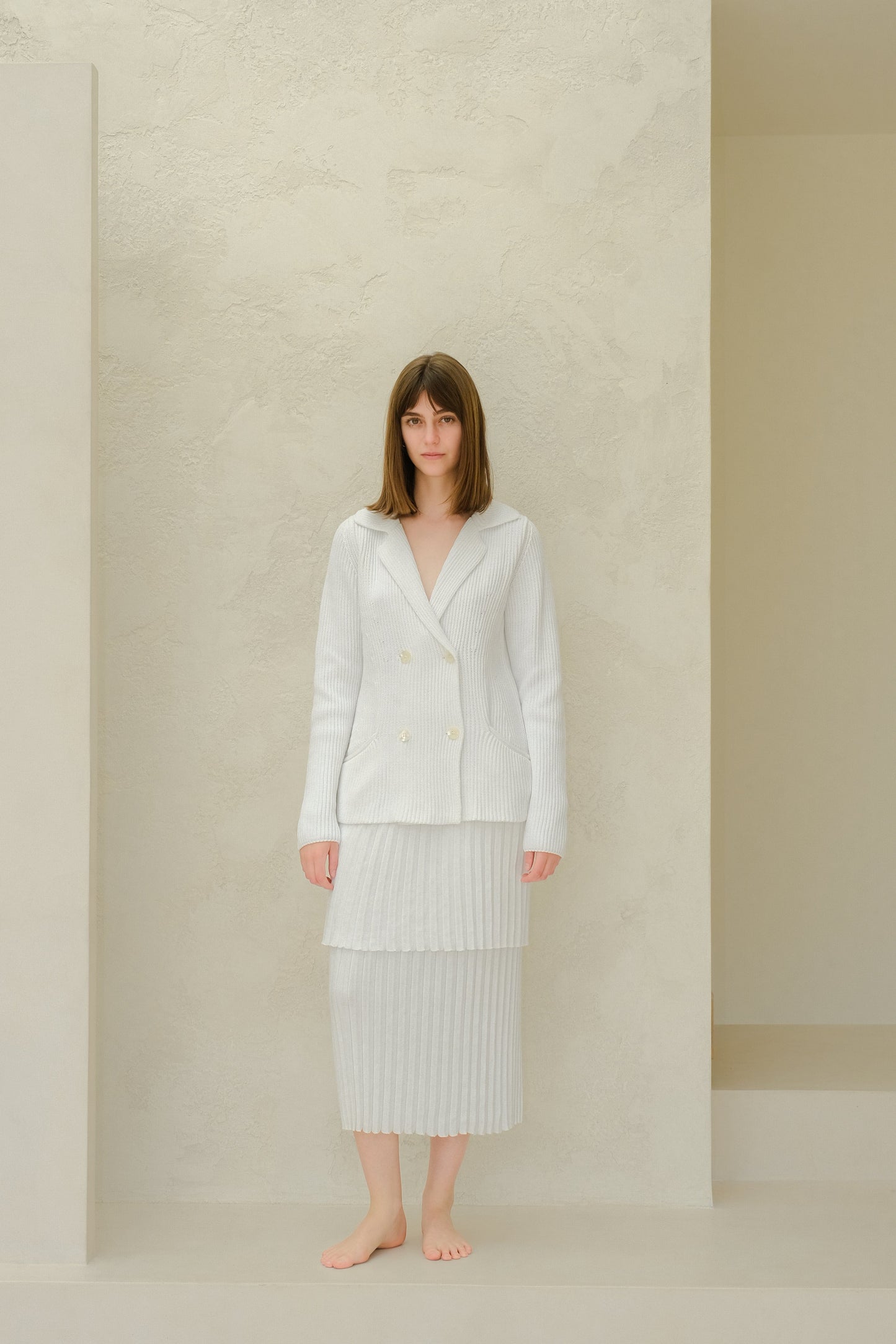 Model wearing a light knit double-breasted blazer and pale pleated long skirt against a neutral studio wall.