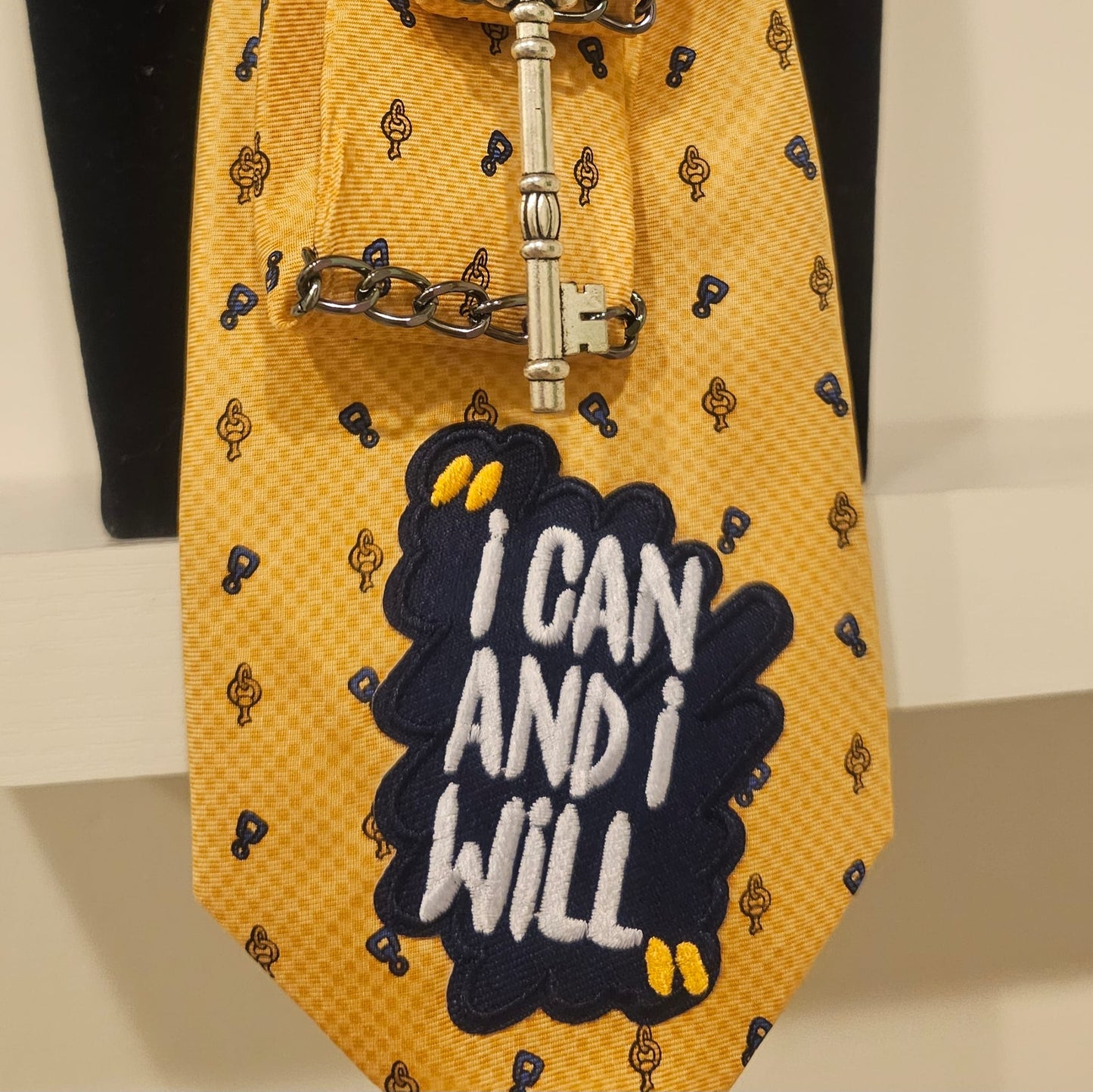 Detail of I can and I will 4-in-1 Jeweled Tie showing the navy embroidered patch I CAN AND I WILL on the yellow tie with metal hardware.