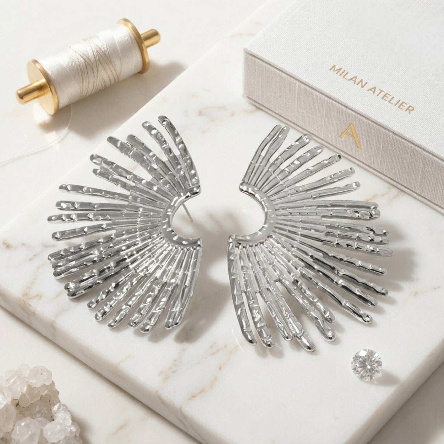 Silver wing-shaped earrings on a marble surface with a spool of thread and a box labeled 'Milan Atelier'.