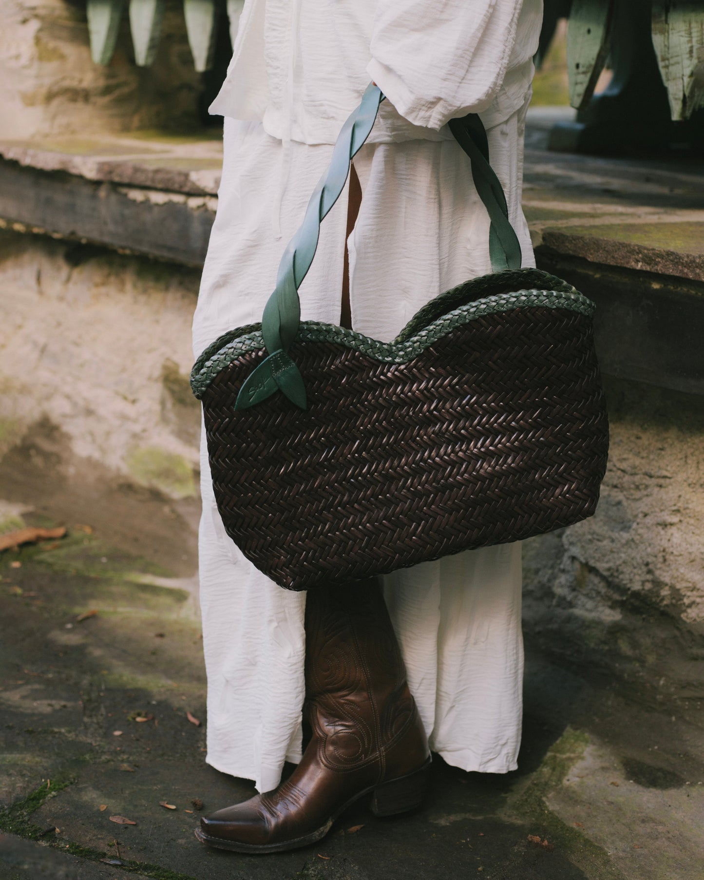 Léafa - Handwoven Leather Shoulder Bag | Green & Coffee featuring woven dark leather body with twisted green leather straps held next to brown boots.