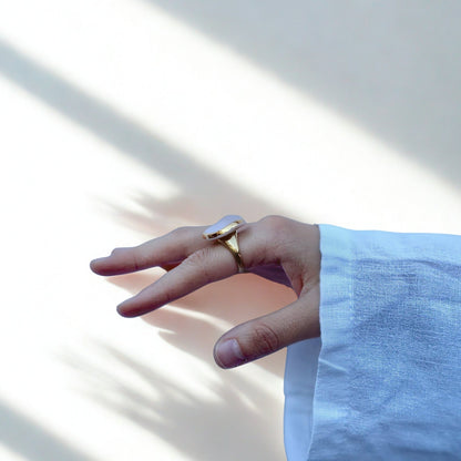 Hand wearing a pink and gold ceramic ring with a white linen sleeve against a neutral background