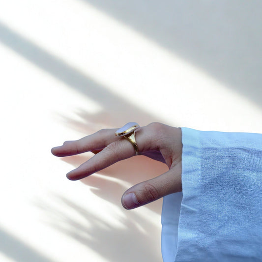 Hand wearing a pink and gold ceramic ring with a white linen sleeve against a neutral background