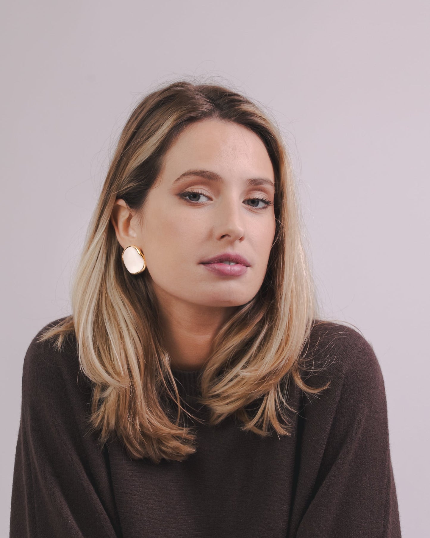 Woman wearing white and gold ceramic earrings with a brown sweater against a plain background