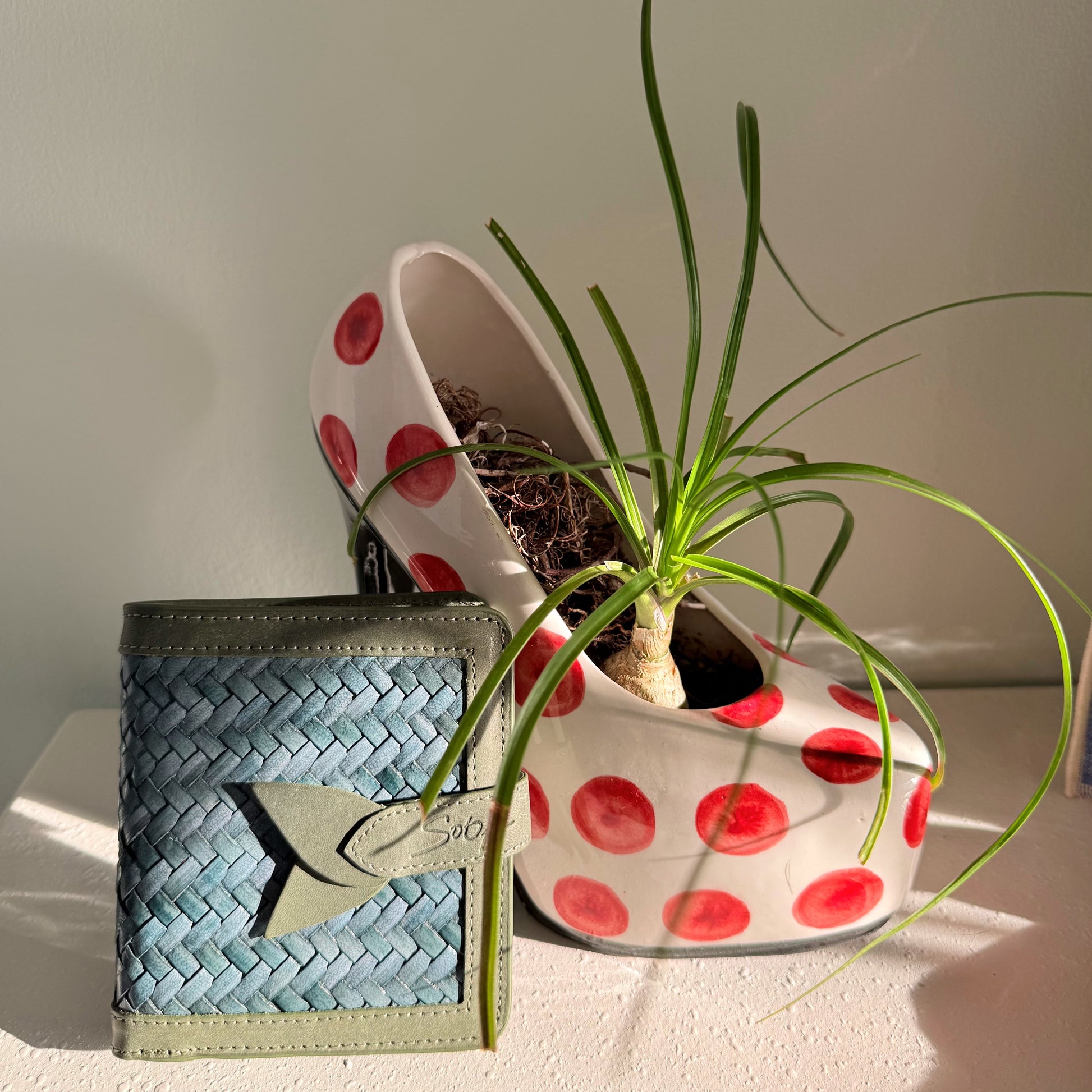 Lumi - Handwoven Leather Wallet | Avocado & Teal showing woven leather pattern with smooth leather trim beside a ceramic pot with green plant.