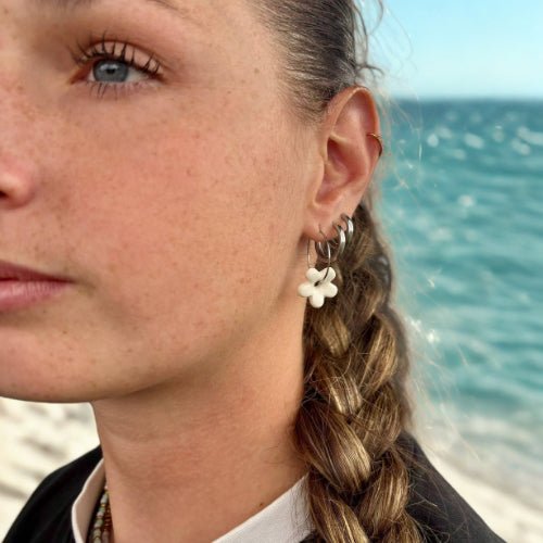 Mimì white floral ceramic dangle earring on a woman's left ear with an ocean backdrop