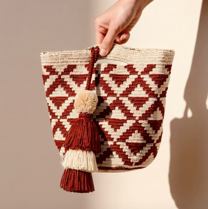Hand holding a woven mochila bag with geometric pattern and tassels against a plain background