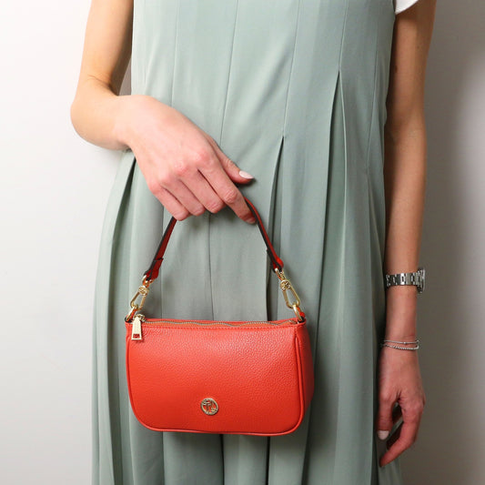 Parfait leather clutch in orange coral with gold hardware and a red strap held by a model in a mint dress