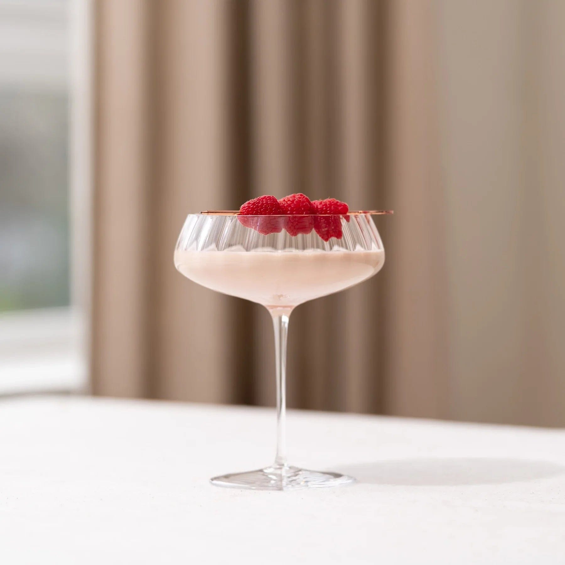 Dusty rose coupe glass with pale pink cocktail and fresh raspberries on the rim