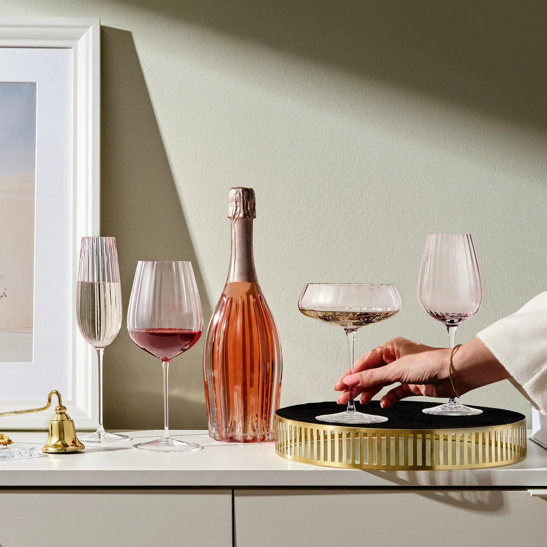 Hand places a dusty rose coupe glass from the Round Up set of two on a round black and gold tray beside a pink champagne bottle