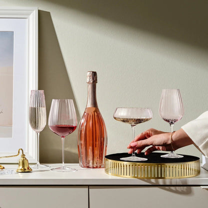 Hand places a dusty rose coupe glass from the Round Up set of two on a round black and gold tray beside a pink champagne bottle