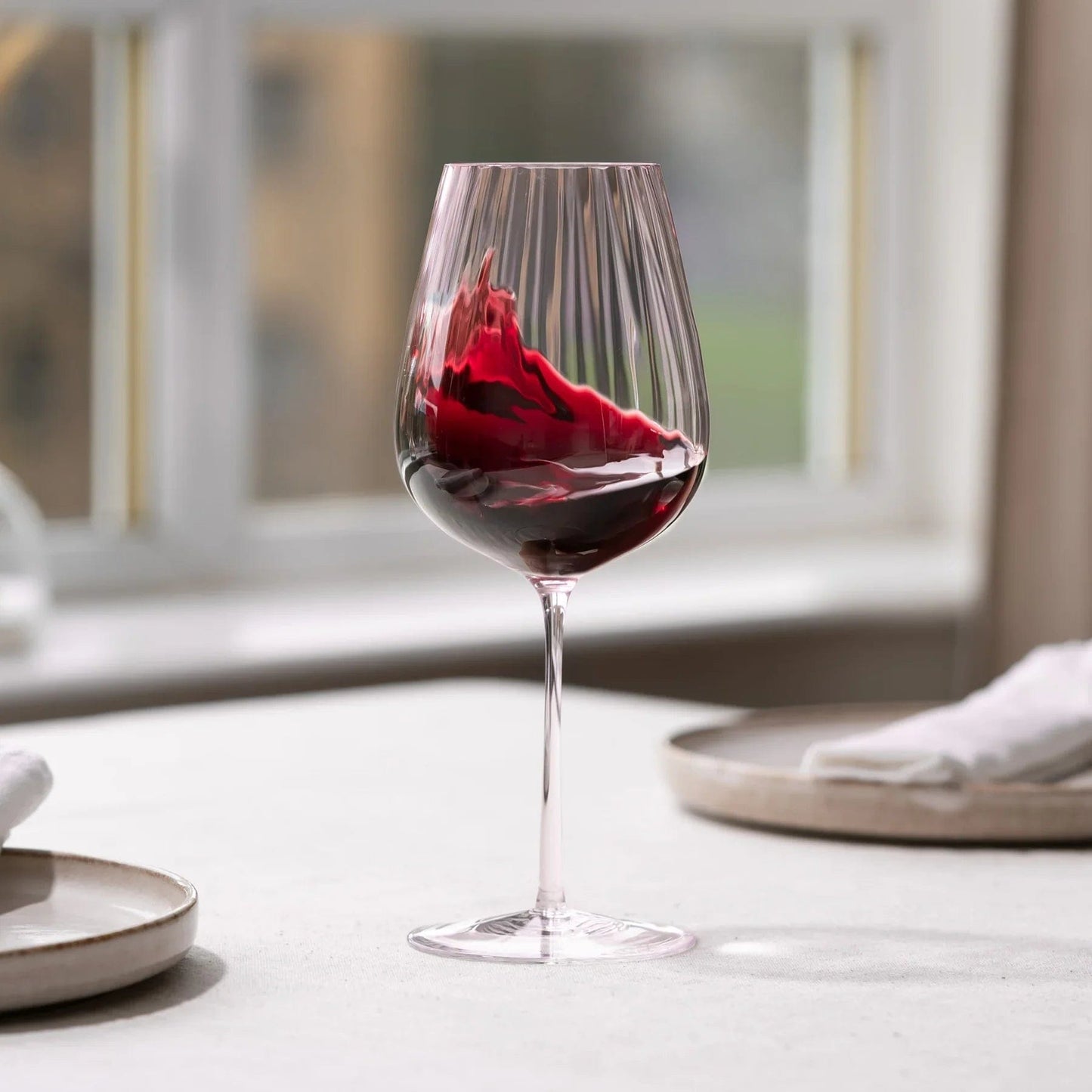 Dusty rose ribbed wine glass with red wine on a dining table near a window