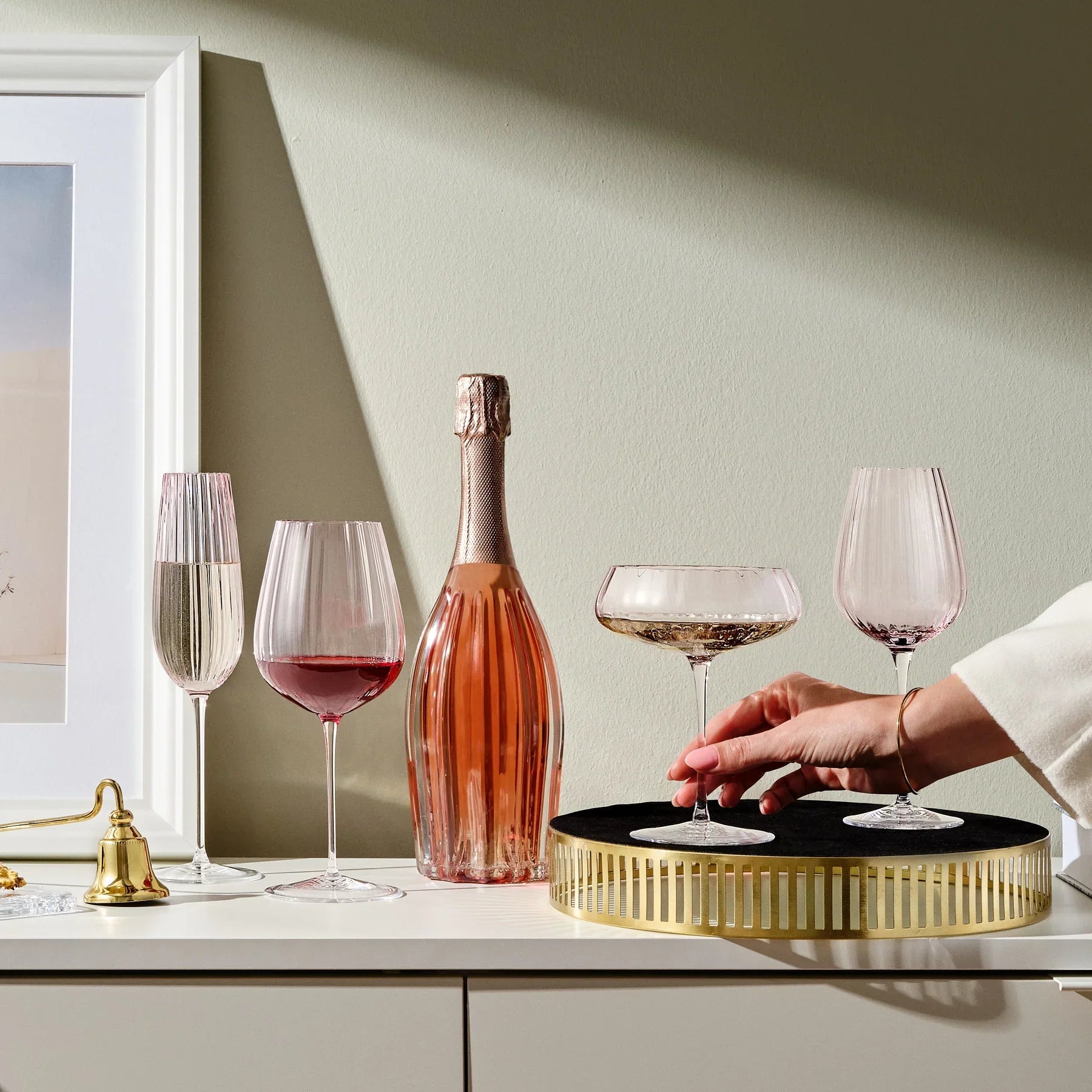 Two dusty rose white wine glasses beside a champagne bottle on a white sideboard