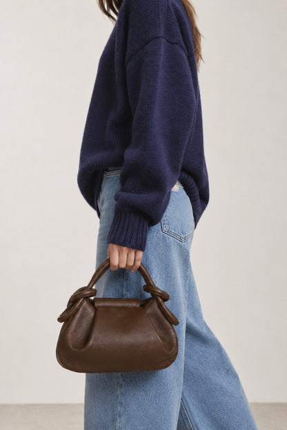 A person wearing a navy knit sweater and light blue jeans carries a small brown leather handbag with top handles against a plain light background.