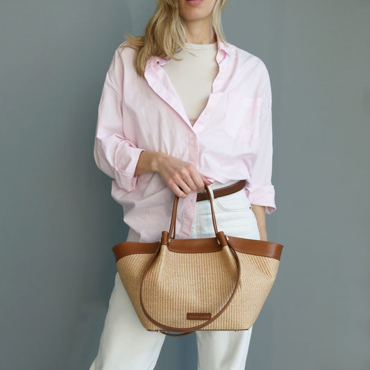 Model in a pale pink shirt holds a Soleil raffia shopping bag with brown leather trim.