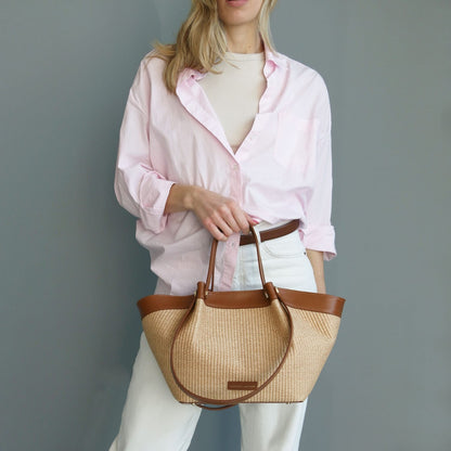 Model in a pale pink shirt holds a Soleil raffia shopping bag with brown leather trim.