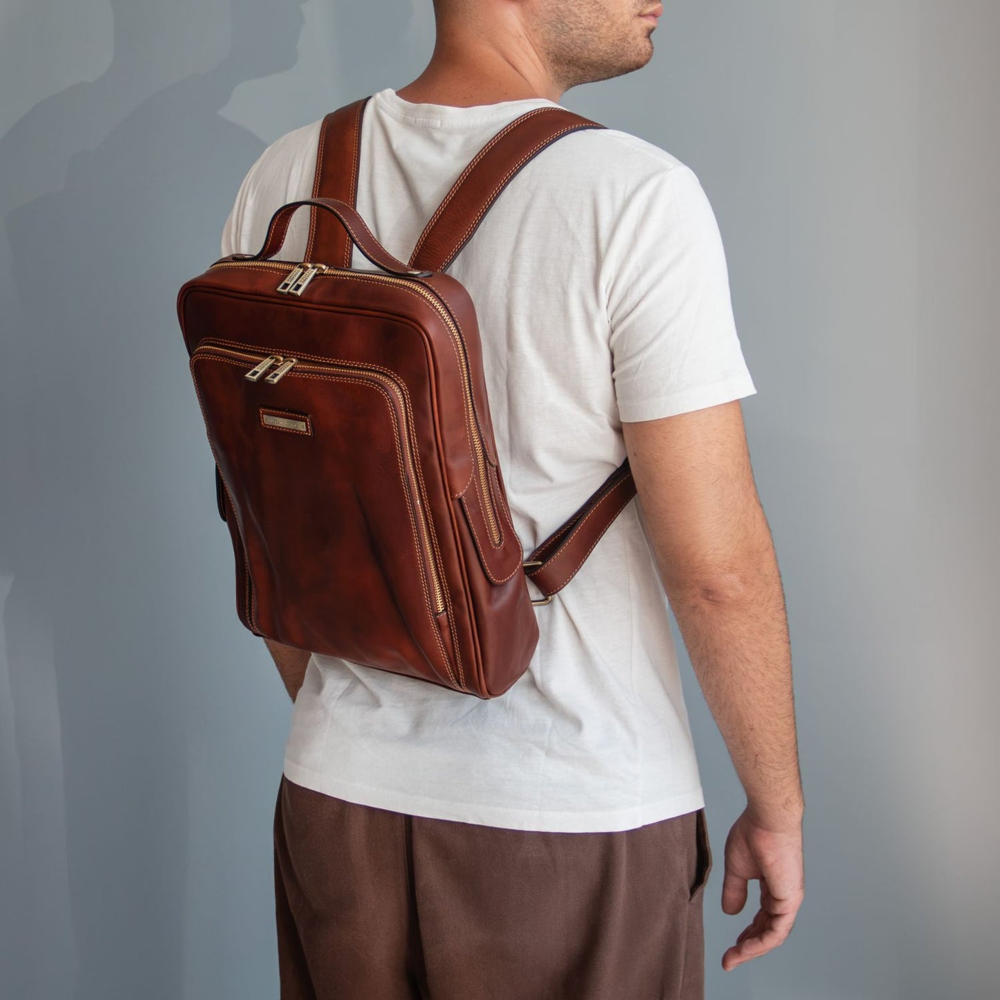 A man wearing a white t-shirt and brown pants, shown from the back and side, carrying a brown leather laptop backpack with gold-tone hardware against a gray studio background.