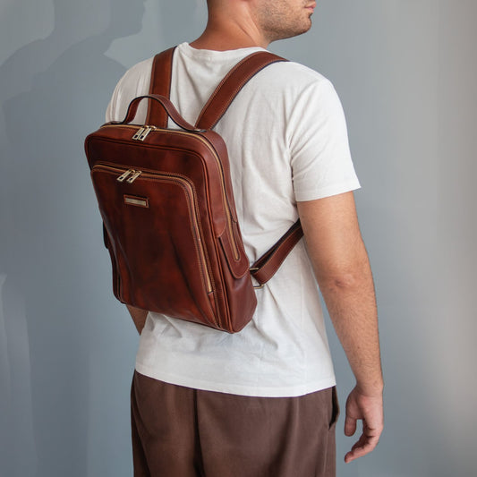 A man wearing a white t-shirt and brown pants, shown from the back and side, carrying a brown leather laptop backpack with gold-tone hardware against a gray studio background.