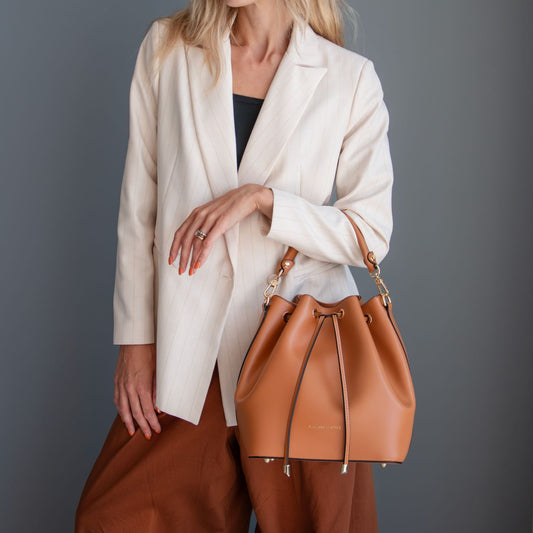 A model wearing a cream blazer and brown trousers carries a tan leather drawstring bucket bag with gold-tone hardware against a dark gray studio background.