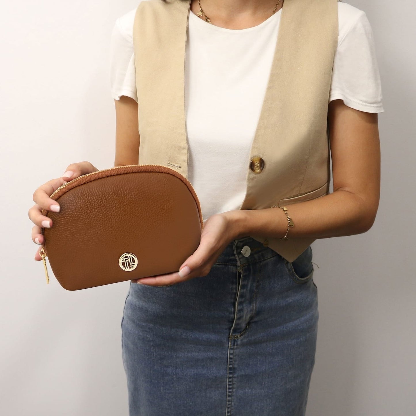 Rounded tan brown soft leather toiletry case with gold zipper held by a person in a white top.